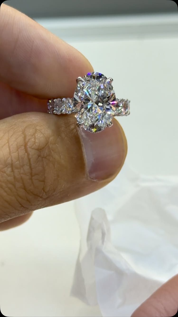 Oval-cut diamond engagement ring held between two fingers against a white background