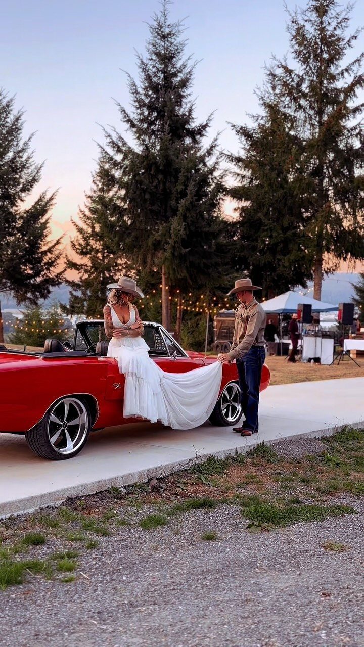 Bride in white wedding dress and cowboy hat sitting on red convertible car