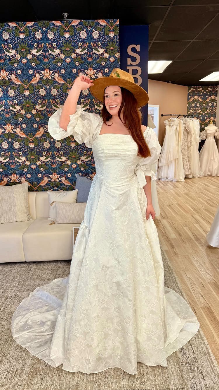 Woman in a white puff-sleeve wedding dress and brown wide-brimmed hat in bridal shop.