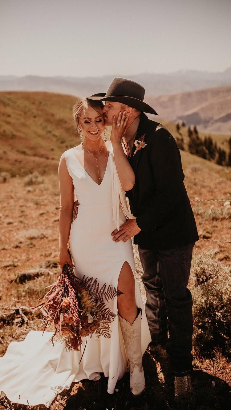 Bride in white wedding dress and cowboy boots embracing groom in western attire.