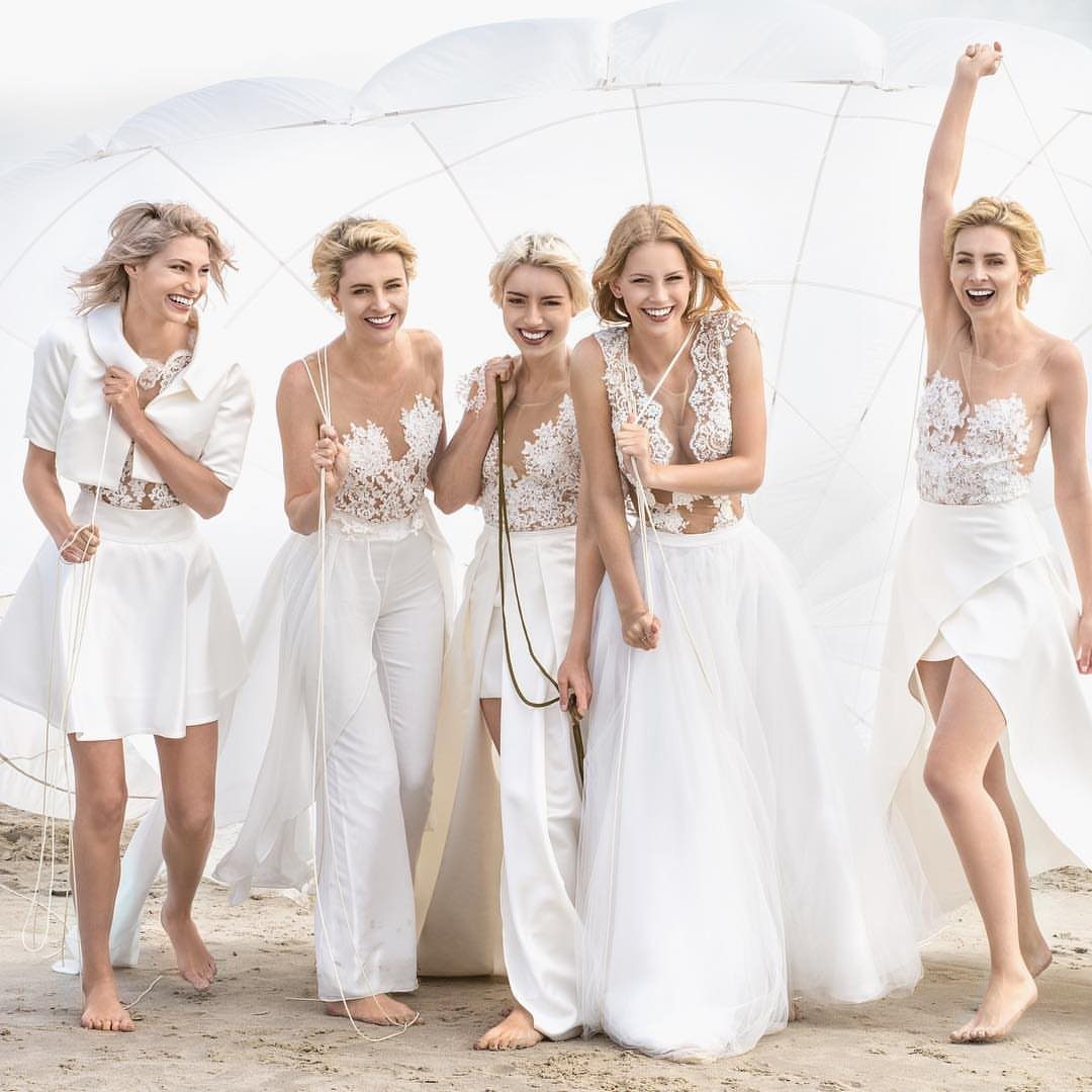 Five women in white bohemian two-piece bridal outfits smiling on a sandy beach.